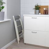 Kleiner weißer Küchentisch mit vier Schubladen, grau gepolsterten Klappstühlen, Holztheke mit Griff, grau geflieste Wand, Holzboden.