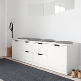 Minimalistische weiße NORDLI-Truhe mit gepolsterten Sitzen, vier runden grauen Hockern, die mit einem schwarz-grauen Teppich und einem einzelnen hellbraunen Stiefel kontrastieren.