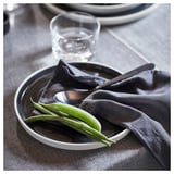 Dunkler Teller mit grünen Zuckerschoten und Löffel. Glas und Serviette daneben. Modernes, minimalistisches Design.