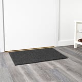 Dunkelgraue rechteckige Fußmatte, die an einer weißen Wand auf einem Holzboden platziert ist und für den Außenbereich konzipiert ist.