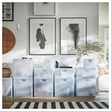 Durchsichtige Aufbewahrungsboxen aus Kunststoff mit gestapelten Deckeln, die zum Organisieren von Gegenständen konzipiert sind, unter gerahmter Wandkunst auf einem schwarz-weiß gestreiften Teppich platziert.