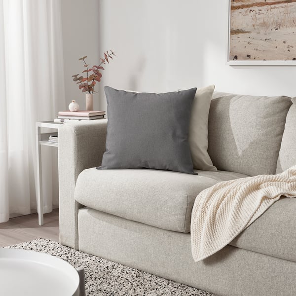 PARADISBUSKE Kissen, grau, weiche Struktur, quadratische Form. Großformatig, sitzt auf einem Sofa, in der Nähe einer Vase.