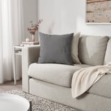 PARADISBUSKE Kissen, grau, weiche Struktur, quadratische Form. Großformatig, sitzt auf einem Sofa, in der Nähe einer Vase.