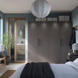 Modernes Schlafzimmer mit grauem PAX-Kleiderschrank, weißer Bettwäsche, blauem Mantel, Pflanze und Pendelleuchte.