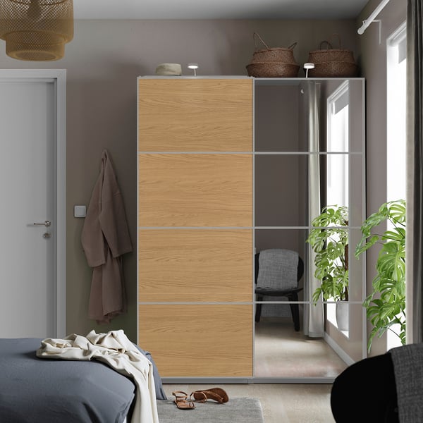 Modernes Schlafzimmer mit hellem Holzschrank, Spiegelfinish, weißer Tür, grauer Bettwäsche, Körben und Pflanzen.