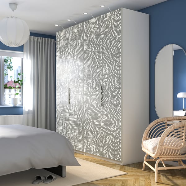 Modernes Schlafzimmer mit großem weißem PAX-Kleiderschrank, Bett, Korbstuhl, Lampe, Spiegel und Pflanze auf der Fensterbank.