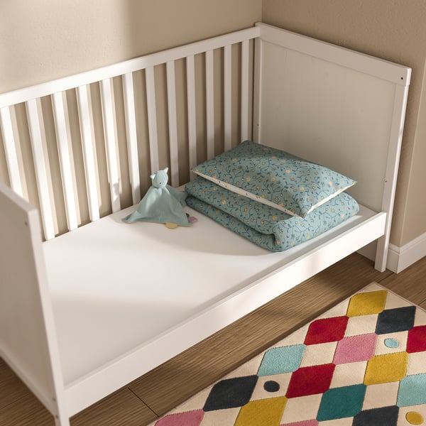 Eine weiße PELLEPLUTT Kleinkindmatratze in einem Kinderbett mit einem blau gemusterten Kissen und einem hellblauen Spielzeug. Das Zimmer verfügt über einen bunten geometrischen Teppich.