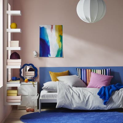 Schlafzimmer mit farbenfrohem Dekor: blauer Bettbezug auf gestreiftem Kopfteil, lebendige Kissen, schwebende Regale und abstrakte Malerei.