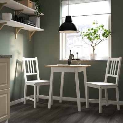 Kleiner Schreibtisch und Stühle aus weißem Kiefernholz in einem kompakten Raum mit grünen Wänden und großen Fenstern, eine stilvolle, minimalistische Einrichtung.