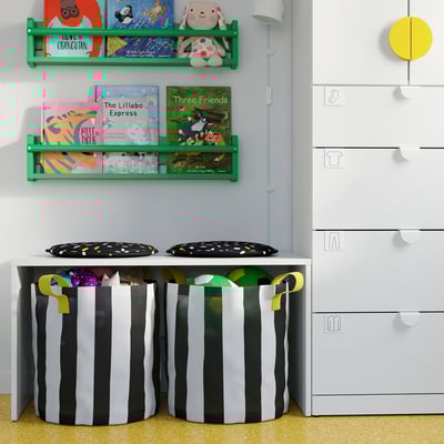 Kinderzimmer mit gestreiften PUGGEHATT-Pads , niedrigem Tisch, grünen Bücherregalen mit Büchern, gelber Lampe, weißen Schubladen.