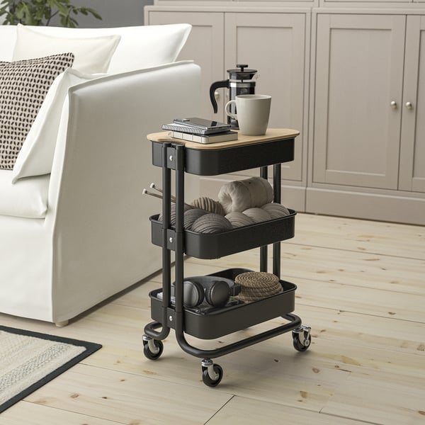 Kleiner schwarzer RÅSKOG-Wagen in der Nähe einer weißen Couch, der unter Tische passt. Fasst Kaffeemaschine, Tassen und Garn. Hergestellt aus Stahl und Bambus.