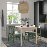 Kleiner heller Esstisch aus Holz mit grünen Stühlen, moderner geflochtener Hängelampe, Teppich, Pflanzen und schwarzen Küchenschränken.