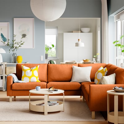 Gemütliches Wohnzimmer mit orangefarbenem SALTSJÖBADEN Ecksofa, gelben Kissen, einem Holzcouchtisch, beigem Teppich, Kunstdrucken und weißer Pendellampe.