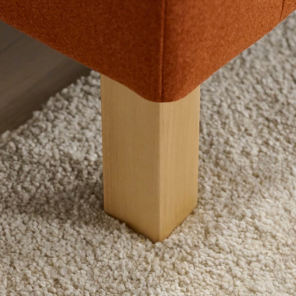 Nahaufnahme des orangefarbenen Sofabeins auf strukturiertem weißem Teppich, das ein stabiles, hochwertiges Design zeigt.