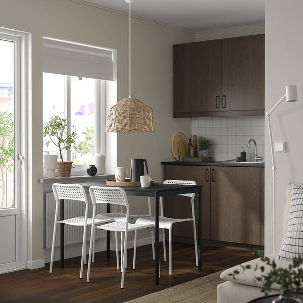 Gemütliche Küche mit weißen Sansberg-Stapelstühlen, Holztisch und Pendelleuchte aus Korbgeflecht. Dunkle Holzschränke und großes Fenster mit Pflanzen.