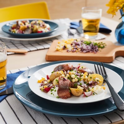 Tisch gedeckt mit Tellern mit mariniertem Hering, Kartoffeln und Beilagen. Biergläser und gelber Stuhl sichtbar.