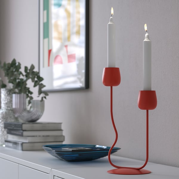 Roter Kerzenständer mit zwei brennenden Kerzen auf einem Regal, neben einer Pflanze und Büchern.