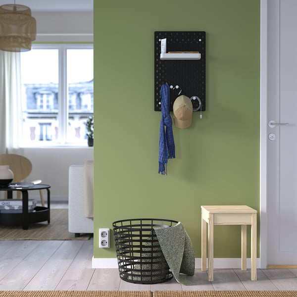 Schwarzes SKÅDIS-Steckbrett mit Accessoires an grüner Wand, neben weißer Tür und Holzhocker, auf dem ein Hut, ein Schal und ein Korb ausgestellt sind.