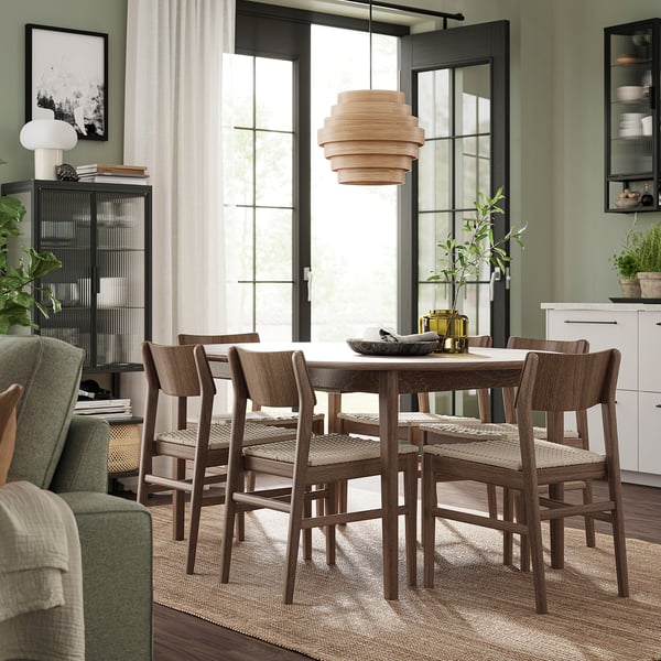 Modernes Esszimmer mit SKANSNÄS Tisch aus dunklem Holz, vier Stühlen, Leuchte und großen Fenstern. Grüne Wände, weiße Vorhänge, Pflanzen und Kunstwerke.