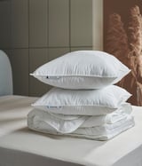 Drei weiße Kissen, ordentlich gestapelt auf einem Bett. Das obere Kissen ist rechteckig, das mittlere quadratisch, das untere ist eine gefaltete Bettdecke. Alle Artikel sind weiß und modern.
