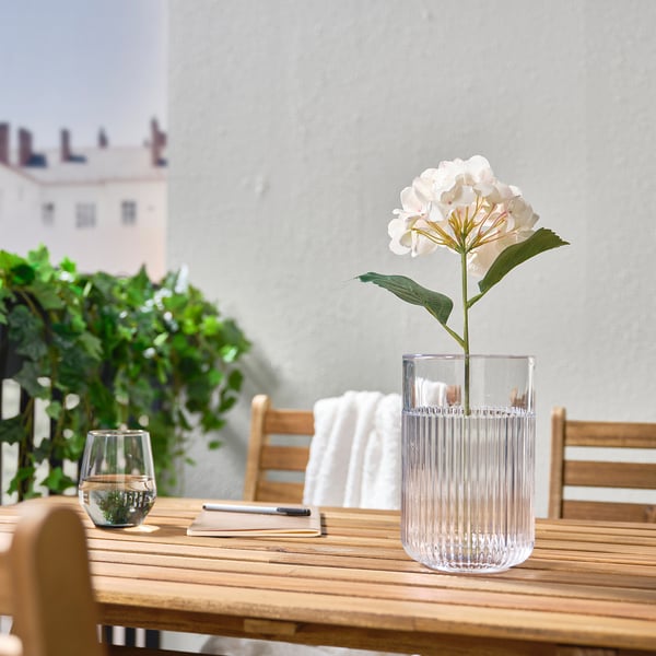 Weiße künstliche Blume in klarer gerippter Vase auf Holztisch, mit Stift und Pflanze.
