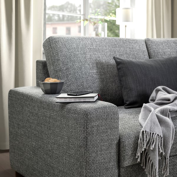 Modernes graues Sofa mit großen Kissen und dunklen Kissen. Schüssel mit Essen, Büchern und Fernbedienung auf der Armlehne. Decke auf der rechten Seite drapiert.