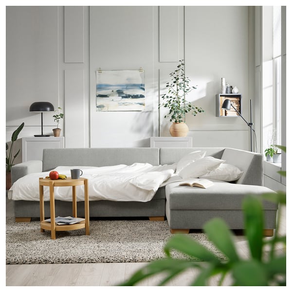 Modernes Wohnzimmer mit hellgrauem Schlafsofa von SÖRVALLEN, weißer Bettwäsche, Beistelltisch aus Holz, großen Pflanzen und abstrakter Wandkunst.