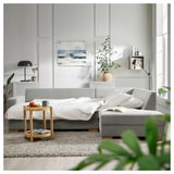 Modernes Wohnzimmer mit hellgrauem Schlafsofa von SÖRVALLEN, weißer Bettwäsche, Beistelltisch aus Holz, großen Pflanzen und abstrakter Wandkunst.