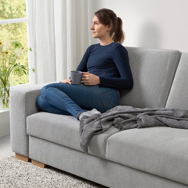 Die Frau sitzt entspannt auf einem grauen SÖRVALLEN-Sofa, hält eine Tasse in der Hand, neben ihr liegt eine kuschelige Decke.