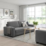 Modernes Wohnzimmer mit grauem SÖRVALLEN-Sofa, passender Ottomane, Couchtisch aus Holz, weißen Vorhängen und großen Fenstern mit Blick ins Grüne.
