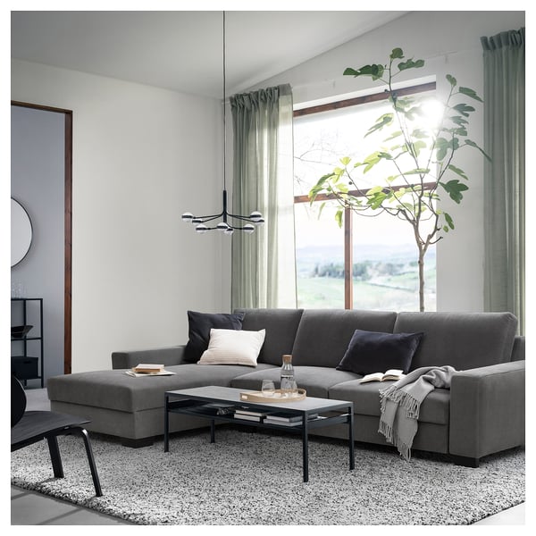 Modernes Wohnzimmer mit einem dunkelgrauen SÖRVALLEN-Sofa, einem schwarzen Couchtisch, großen Fenstern und einer hohen grünen Pflanze.