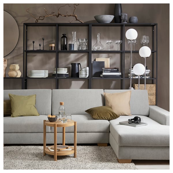 Modernes, minimalistisches Wohnzimmer mit grauem SÖRVALLEN-Sofa, Beistelltisch aus Holz und schwarzen Metallregalen, die mit verschiedenen Einrichtungsgegenständen dekoriert sind.