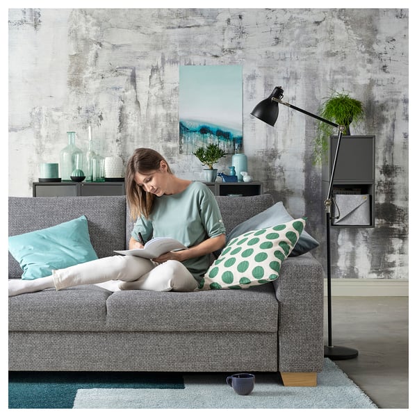 Entspannte Frau, die auf einem grauen SÖRVALLEN-Sofa liest, mit pastellfarbenen Kissen, neben einer schwarzen Stehlampe, vor einer strukturierten Wand mit Kunstwerken und Pflanzen.