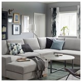 Modernes Wohnzimmer mit grauem SÖRVALLEN-Sofa, Kissen, Couchtisch, Lampe, Bücherregal und großen Fenstern.