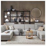 Modernes graues Sektionssofa mit Kissen, Beistelltisch aus Holz, Bücherregal, Teppich und Dekorationsgegenständen in einer gemütlichen Wohnzimmereinrichtung.