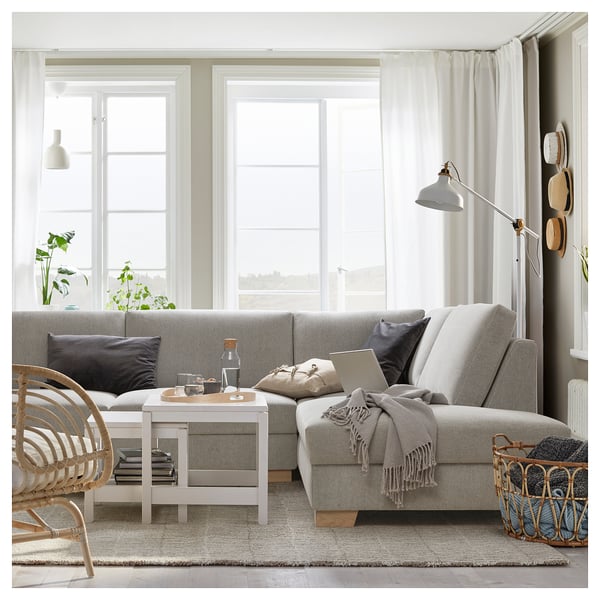 Gemütliches hellgraues Sofa mit mehreren Kissen und weißem Couchtisch in einem hellen Raum mit großen Fenstern und einem Bodenkorb mit Decken.