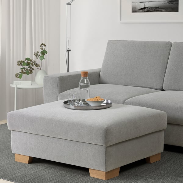 Grauer SÖRVALLEN Fußhocker mit Snacks, Getränken, neben Sofa und Blumen.