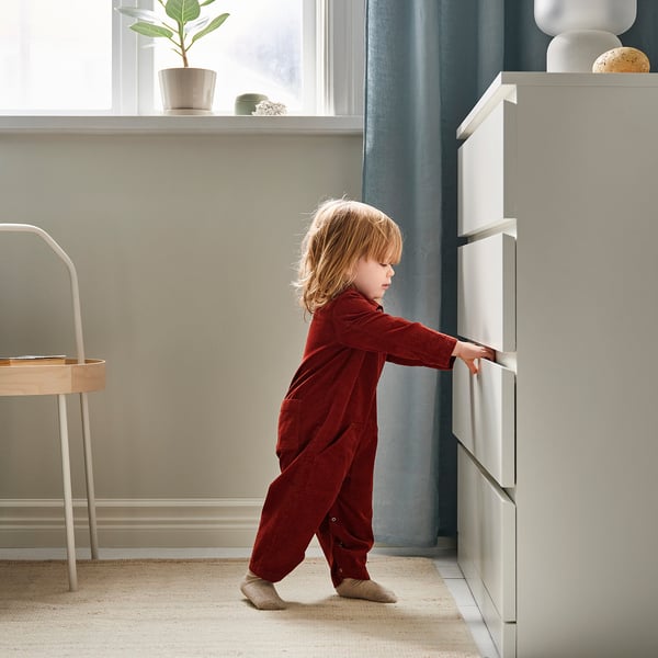 Ein kleines Kind im roten Pyjama steht neben einem weißen Schubladenschrank und greift nach dem Griff. Der Raum ist in neutralen Tönen gehalten und ein blauer Vorhang.