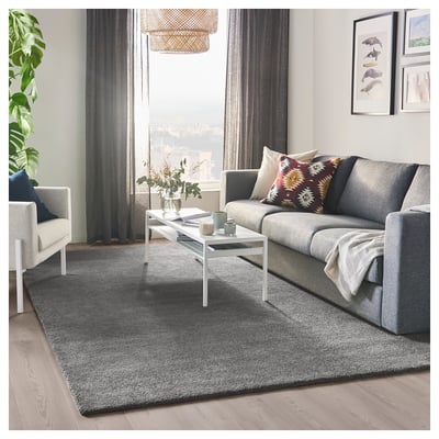Stilvolles Wohnzimmer mit grauem STOENSE-Teppich, grauem Sofa, bunten Kissen, weißem Couchtisch, großem Fenster, grauen Vorhängen und Pflanze.