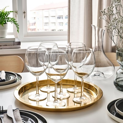 Sechs klare Weingläser auf einem goldenen Tablett neben Tellern und Besteck, in der Nähe eines Fensters mit weißen Vorhängen.