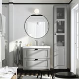 Graues Badezimmer, runder Spiegel, Waschtisch TÄNNFORSEN, weißes Waschbecken, graues Handtuch, Hochschrank, schwarze Akzente.