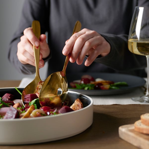 Person mit messingfarbenem Servierlöffel, um Essen aus weißer Schüssel zu servieren. Weinglas neben der Schale.