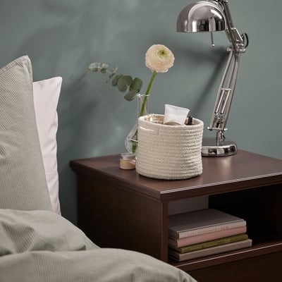Nachttisch mit Lampe, Blumen, Korb und Büchern.