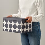 Person, die eine TJENA-Aufbewahrungsbox mit geometrischem Muster hält. Die Box hat einen Griff für einen einfachen Zugriff.