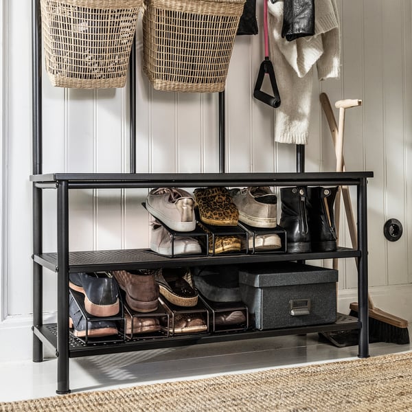 Schuhregal aus schwarzem Metall mit zwei Regalen, in dem verschiedene Turnschuhe und Stiefel aufbewahrt werden können. Positioniert an einer hellgrauen Wand mit Hängekörben und Kleiderhaken.