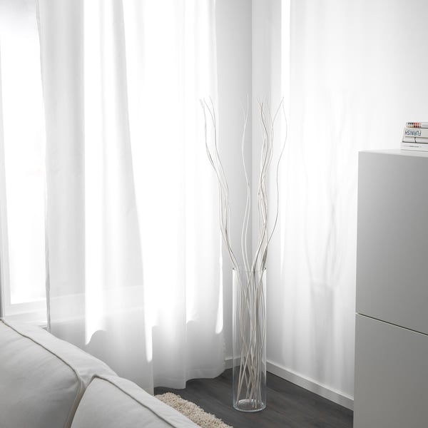 Durchsichtige Vase mit weißen getrockneten Zweigen in der Nähe eines Fensters mit weißen Vorhängen und einer teilweise sichtbaren weißen Couch.