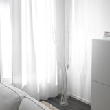 Durchsichtige Vase mit weißen getrockneten Zweigen in der Nähe eines Fensters mit weißen Vorhängen und einer teilweise sichtbaren weißen Couch.
