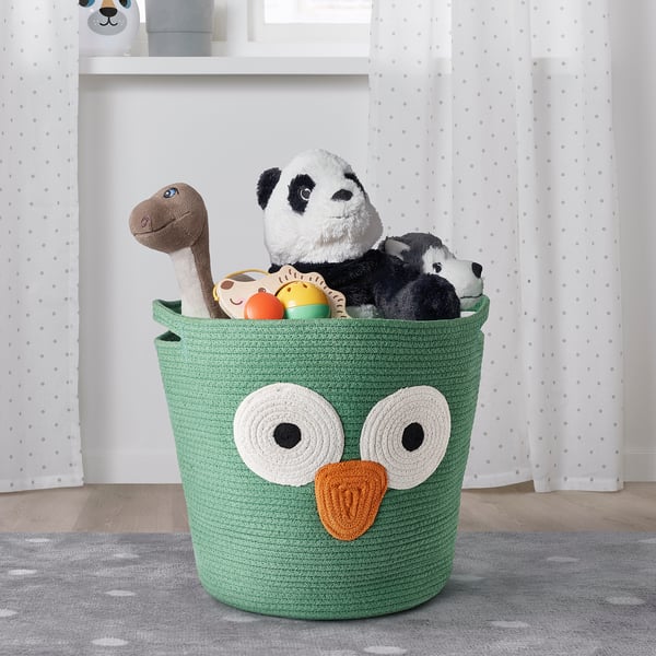 Ein grüner Baumwollkorb mit Eulen-Design hält Plüschtiere auf einem grauen Tupfenteppich.