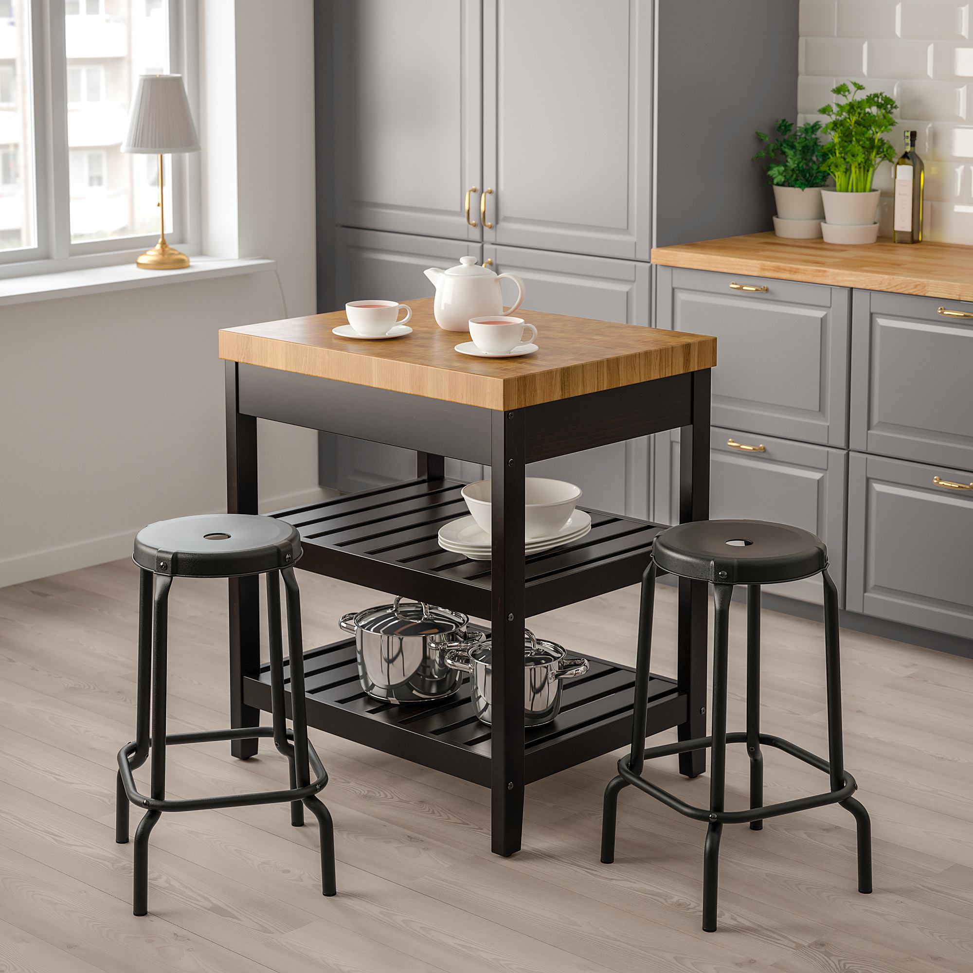 Kompakte VADHOLMA Kücheninsel in Schwarz & Holz. Zwei Hocker, ein Regal mit Kochgeschirr und eine Platte mit Teekanne und Tassen. Passt gut in moderne Küchen.