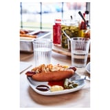 Teller mit Hot Dog und Gewürzen, Gläsern und Flaschen auf Holztisch.